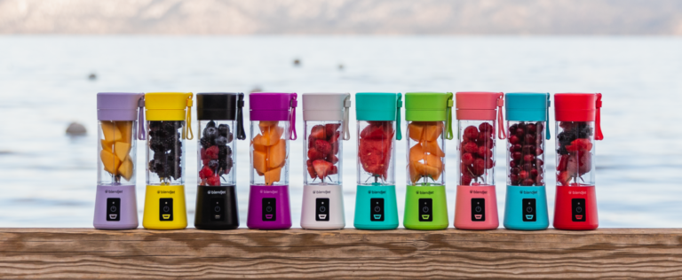 Row of blenders with fruits inside of them, in all different colors, sitting on a deck outside
