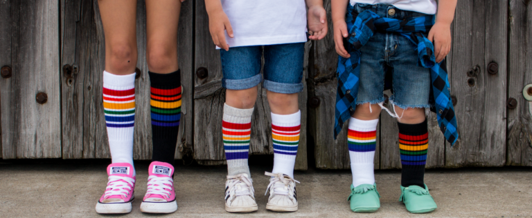 Three kids legs and feet, wearing socks with rainbow strips on them