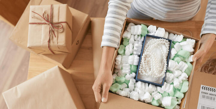 Woman packs a necklace into a larger shipping box full of packing peanuts.