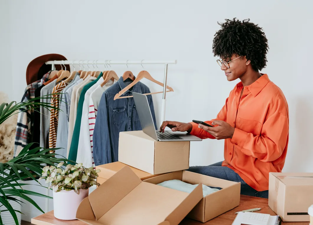 Woman in an orange shirt checking her phone while packing orders into cardboard boxes beside a clothing rack