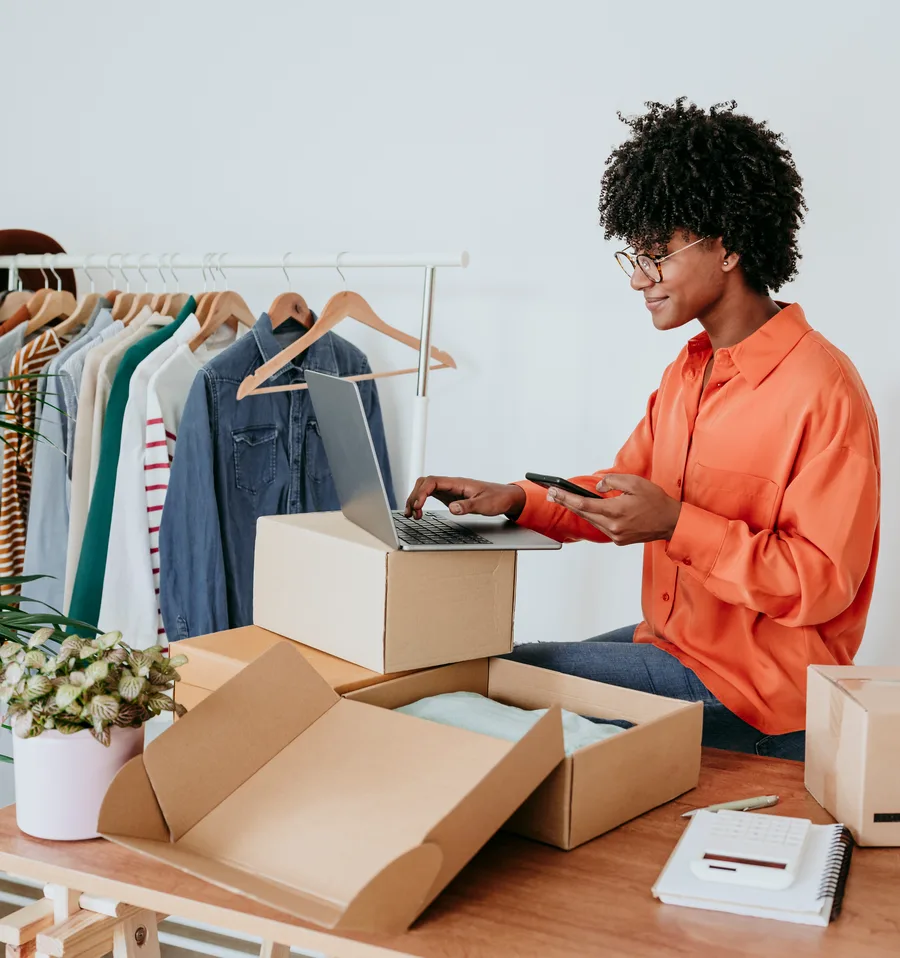 Woman in an orange shirt and glasses typing on a laptop set on a shipping box while holding her phone