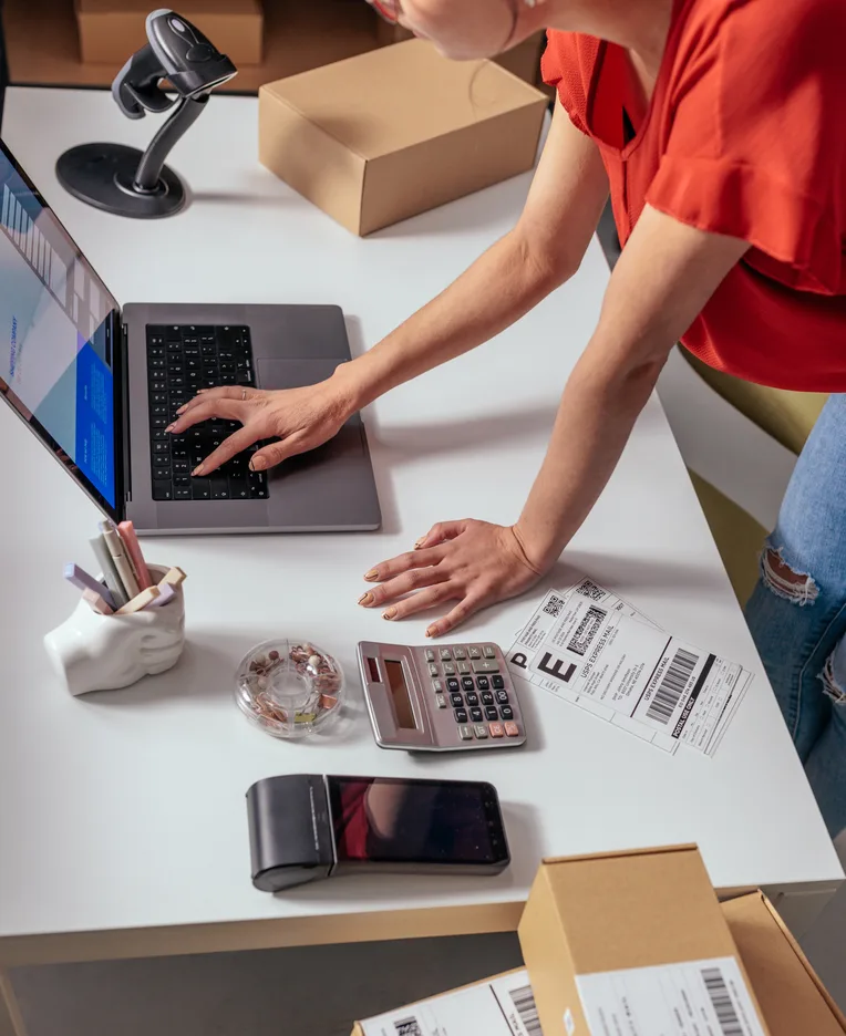 Person typing on a laptop at a packing desk with a barcode scanner, shipping boxes and printed labels