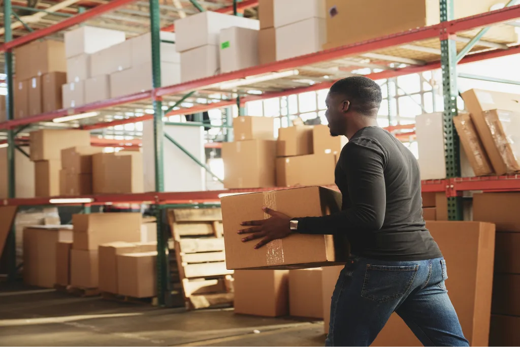 Warehouse worker carrying a cardboard box past tall shelves stacked with packages