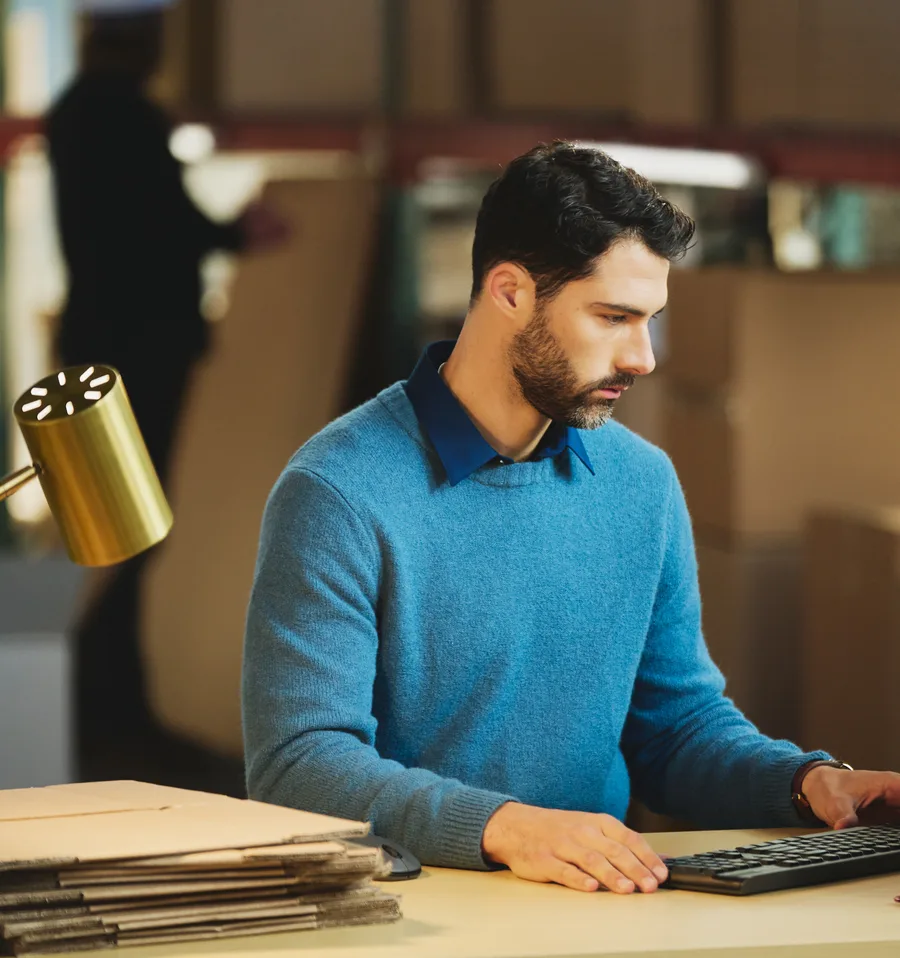 Man in a blue sweater typing on a keyboard at a packing desk in a warehouse