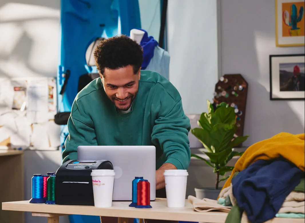 Smiling man in a green sweatshirt working at a laptop beside a label printer and thread spools