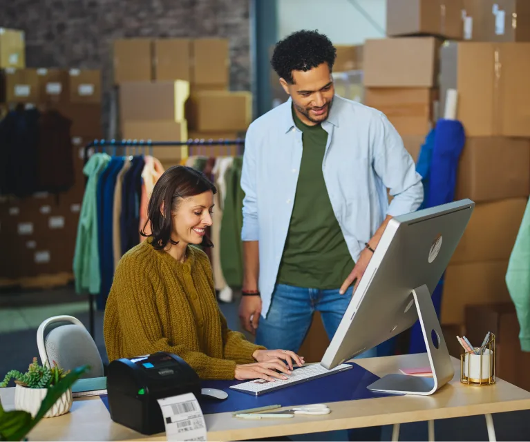 Two coworkers at a computer in an apparel warehouse, one packing orders beside a label printer