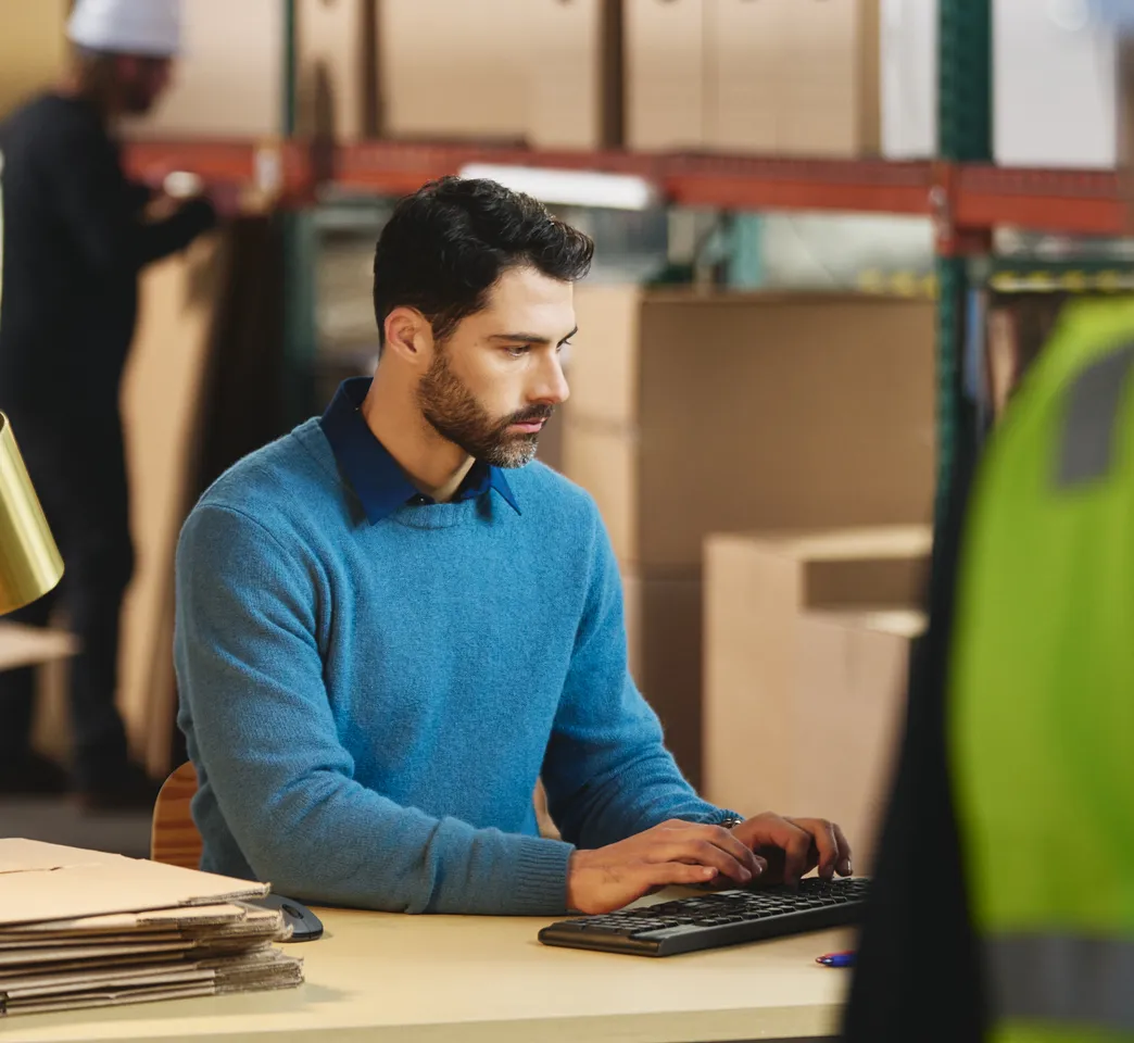 Man in a blue sweater typing at a computer in a warehouse office