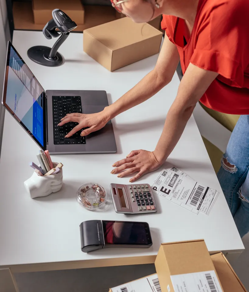 Person at a packing desk typing on a laptop beside a barcode scanner and shipping labels