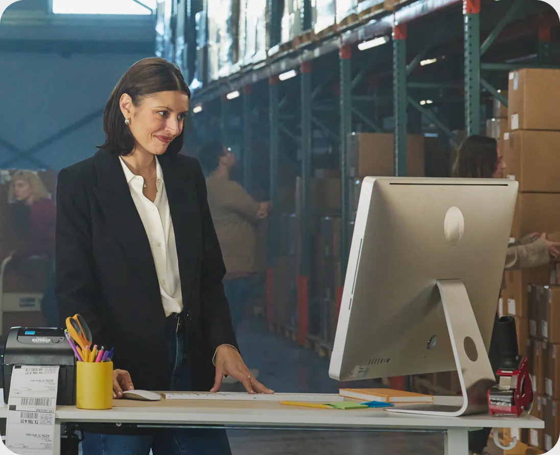 Woman smiling at her computer at a packing desk in a busy warehouse