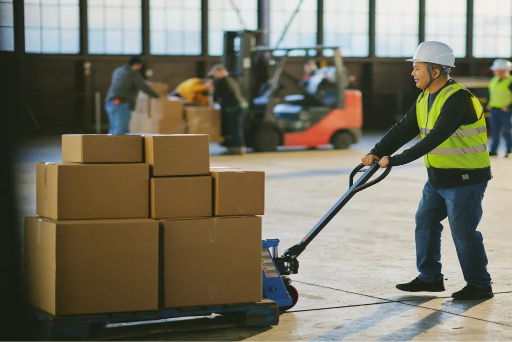 Warehouse worker in a hard hat and safety vest pulling a pallet jack stacked with boxes