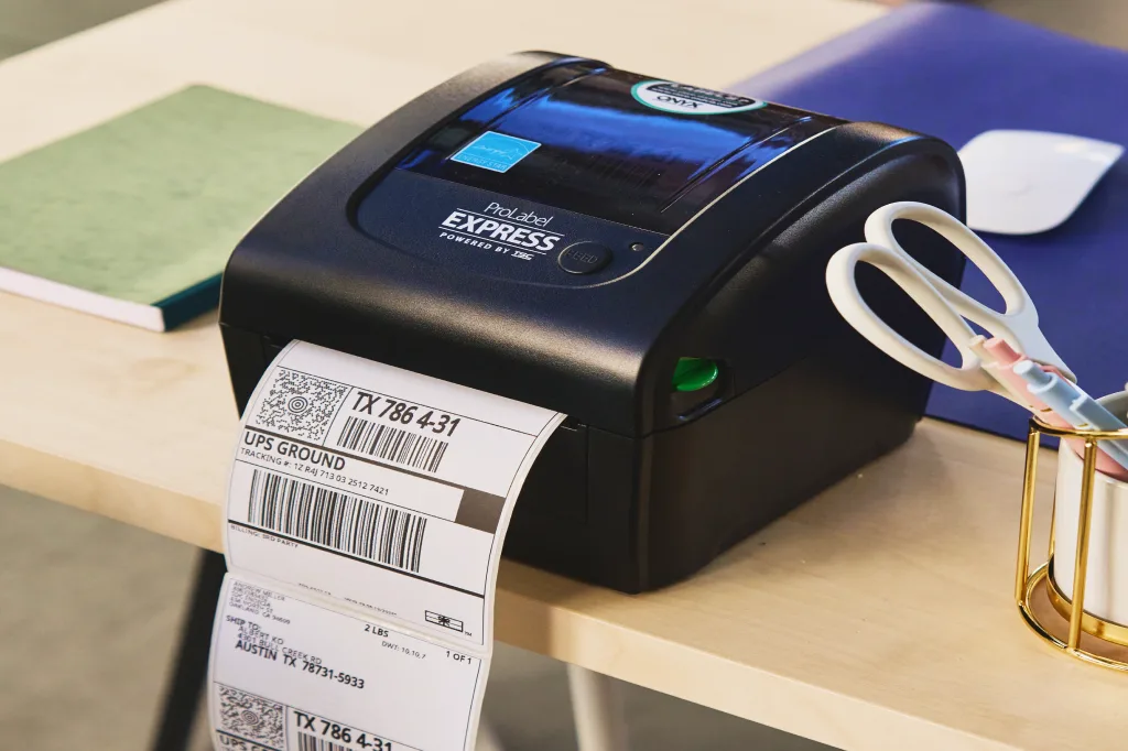 Black thermal label printer on a desk printing a stack of UPS Ground shipping labels, with scissors in a pen cup beside it