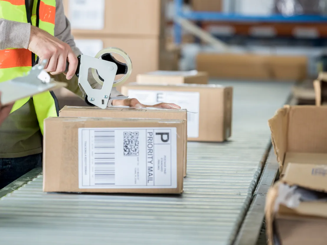 Warehouse worker in a high-visibility vest sealing a Priority Mail box with a tape gun